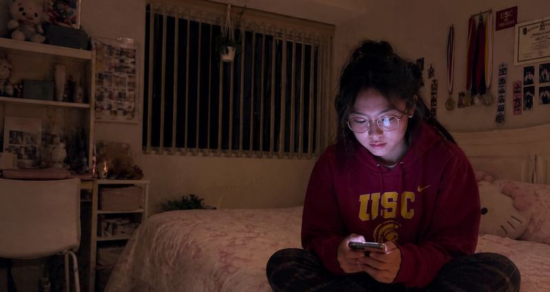 Alicia sits on her bed in a USC sweatshirt, her face lit by her phone.